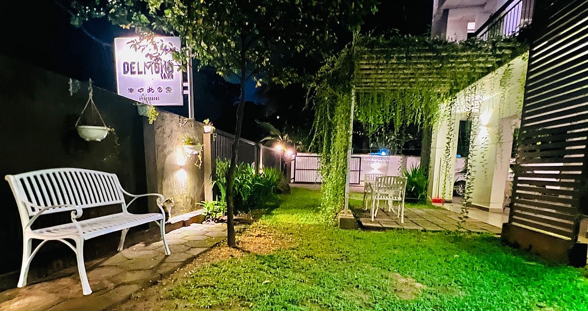 DELMOND Lake garden at dusk with a white bench, hanging lanterns and the illuminated sign