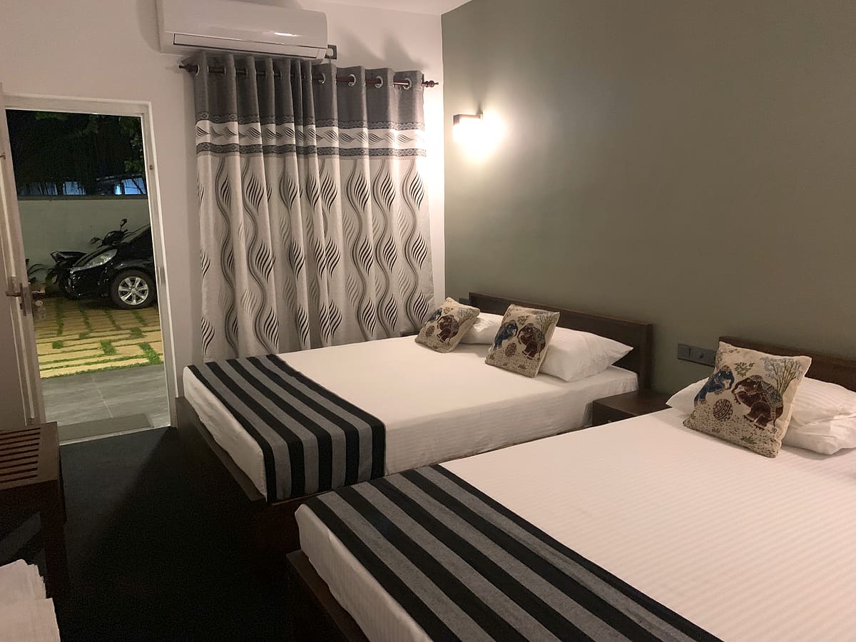Two made-up beds with striped grey runners and floral cushions under an air conditioner, door open to the garden path